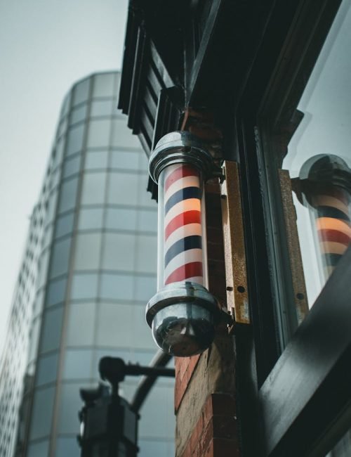 low-angle-view-of-barber-pole-9889982 A traditional barber pole with red, white, and blue stripes stands against a modern city backdrop.